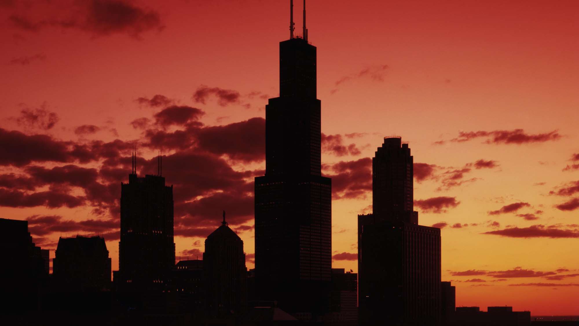 Willis Tower (Sears Tower) silhouette dead-center against a deep red-orange sunset sky, twin antenna masts intact, flanked by the Aon Center and Blue Cross-Blue