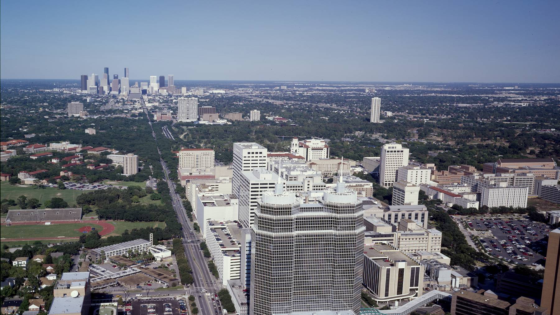 Aerial view of the Texas Medical Center’s twin-towered landmark building with downtown Houston’s skyline visible far in the background down the tree-lined boule