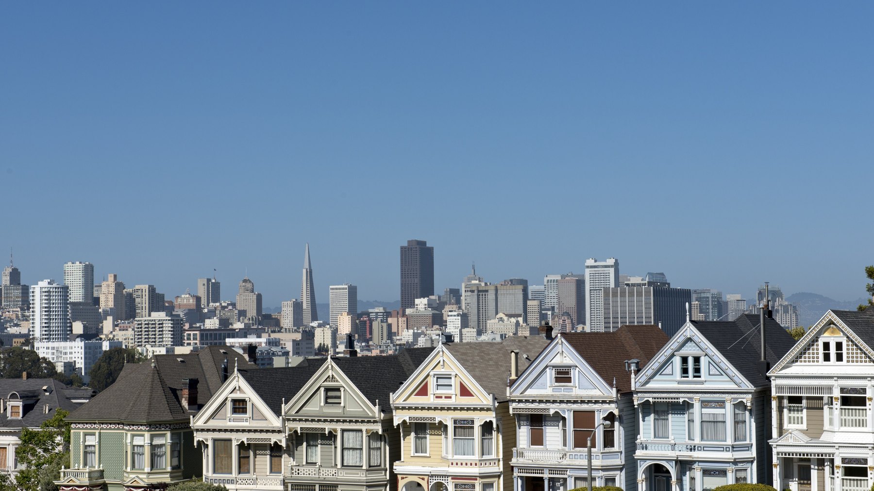 "Painted Ladies" row of Victorian houses on Steiner Street across from Alamo Square Park, downtown SF skyline (Transamerica Pyramid unmistakable) behind, clear